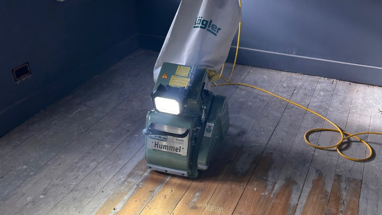 Professional Hummel floor sanding machine in use on old pine floorboards, showing softwood floor restoration in progress inside a dark-painted room.